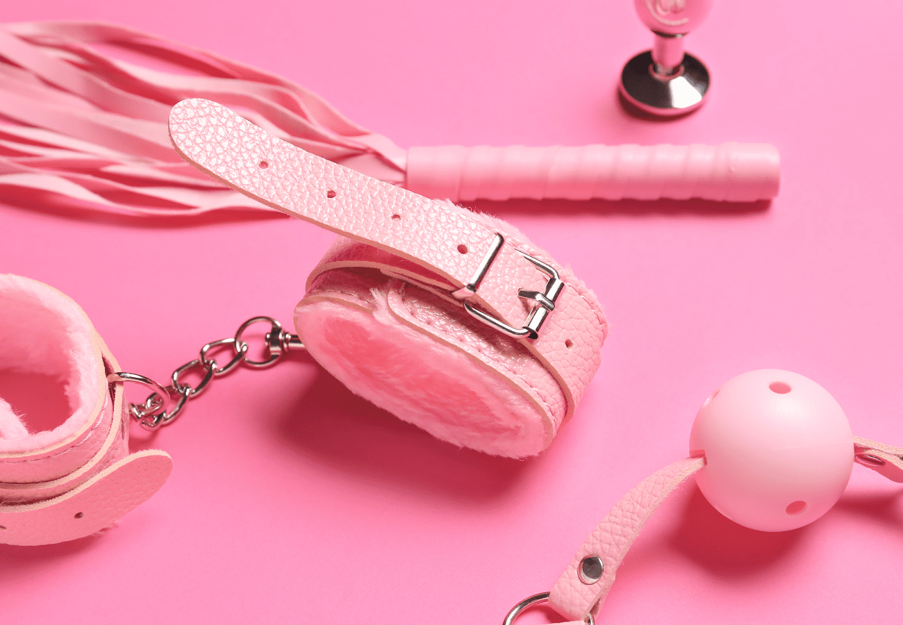 Assorted pink bedroom sex toys like a whip, ball gag, and leather cuffs